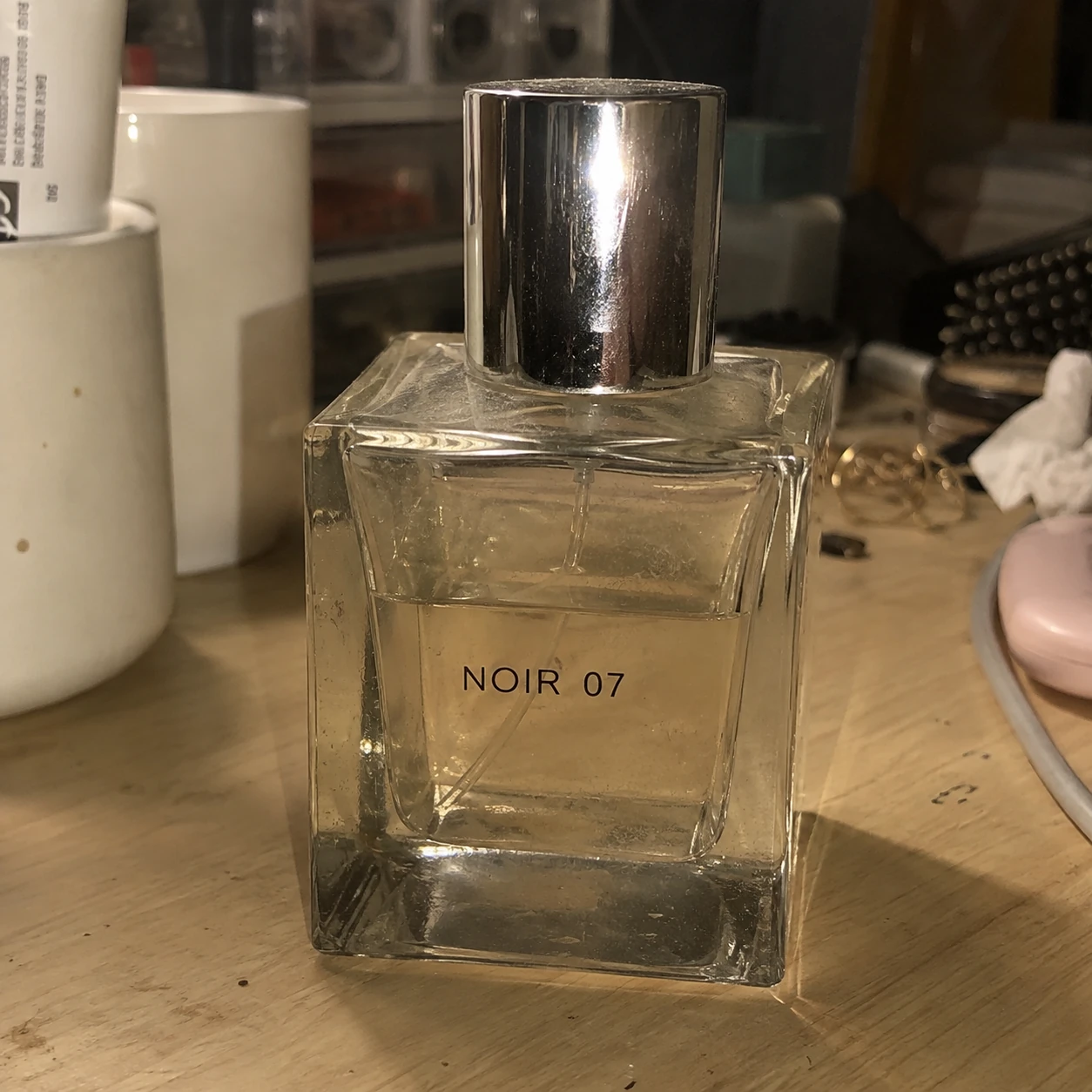 Original product photo: a square glass perfume bottle on a desk with dust, fingerprints, flash glare, a yellow cast and a cluttered background